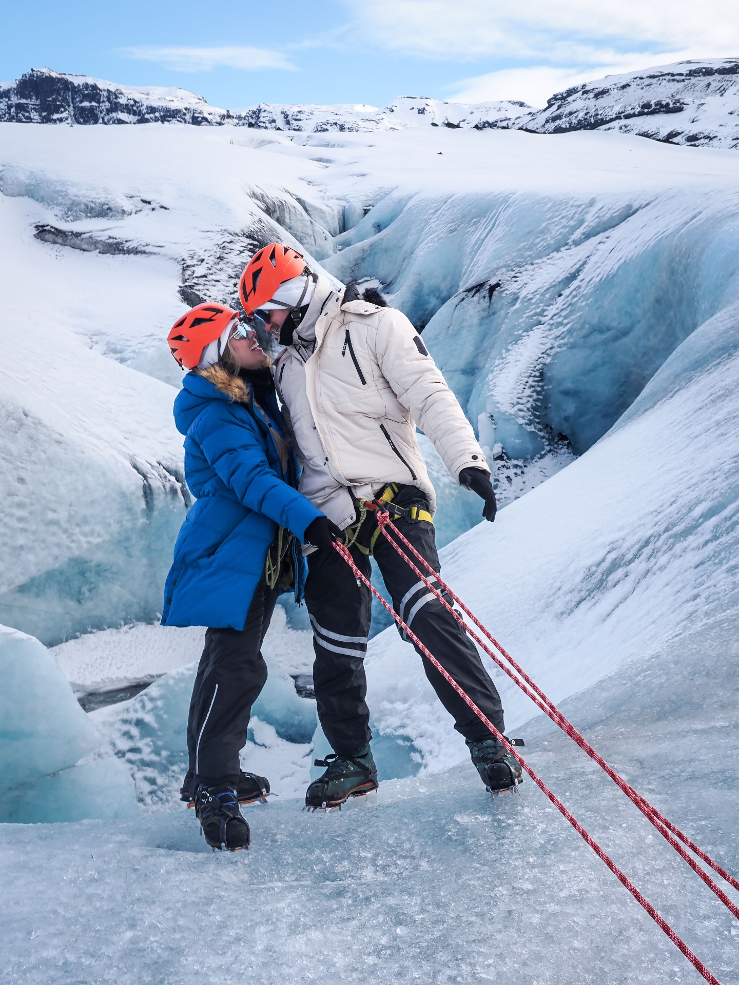 Brittany and Jake in helmets and crampons hanging over the edge of a deep glacier crevasse in Iceland